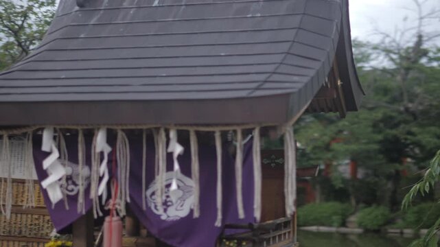 Orbiting shot of a small wooden Shinto shrine decorated with white zigzag paper streamers and a purple cloth curtain on an overcast day in Kyoto, Japan.