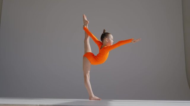 A little Caucasian girl gymnast in an orange leotard demonstrates her skills on a white background. 