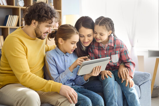 Family parents children using tablet technology for learning. At home on a sofa, two daughters explore education while mom and dad guide. Clear concept of modern family learning and bonding.