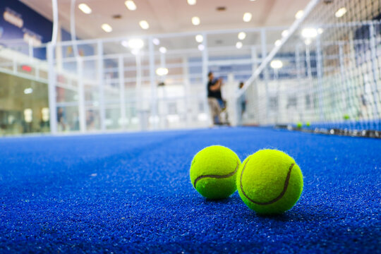 Two bright yellow padel tennis balls rest on a textured blue artificial turf indoor court floor