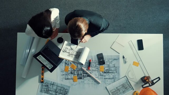 Top view of skilled project manager holding project plan and explain house design to architect engineer at meeting table with equipment, ruler, safety helmet and laptop scatter around. Alimentation.
