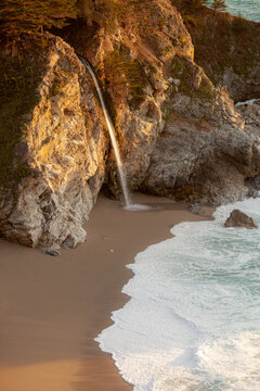 McWay Falls cascades onto a sandy beach in Julia Pfeiffer Burns State Park, Big Sur, California, USA. The golden hour light illuminates the rugged cliffs and the Pacific Ocean waves.
