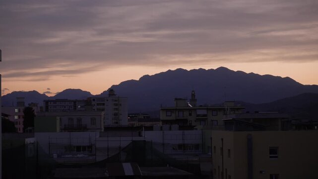 A moody mountain range silhouette looms over urban buildings in Shkoder, Albania, as twilight deepens with soft, fading light and cloudy skies.
