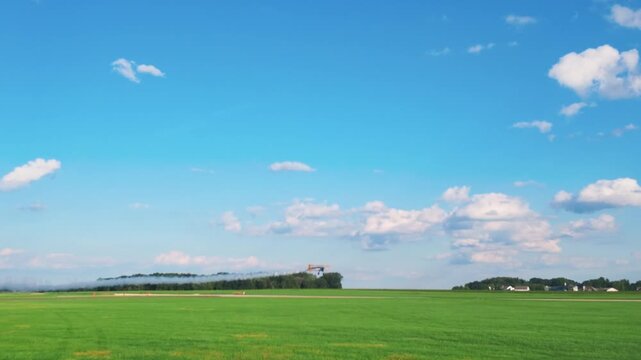 A blue and yellow biplane flying through a bright, cloud filled sky, dispensing a trail of smoke as it flies low near the grass, then into the sun.