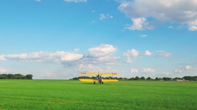 A PT-13 Stearman driving on a bright green grass field under a blue sky of white clouds.