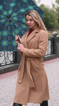 Vertical video. Woman walking. Autumn park. Smiling blonde in beige coat opening umbrella on city path paved stone river embankment.