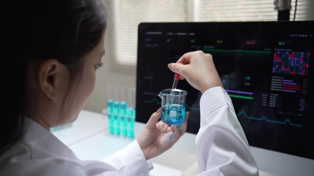 Focused woman scientist conducting chemistry research in laboratory with pipette, blue liquid, glass beaker and computer screen