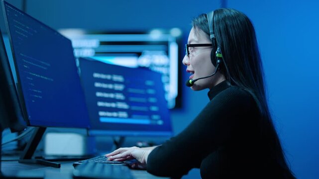 Focused Asian female IT specialist with headset types code on dual monitors in a high tech dark blue server control room. She provides 24 hours, technical support and network cybersecurity monitoring