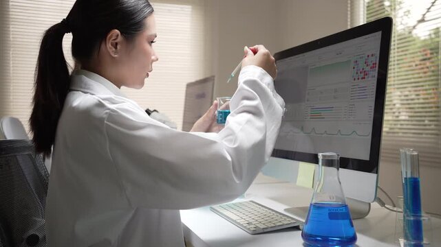 Focused woman scientist in white coat conducts medical research using computer and flask inside laboratory