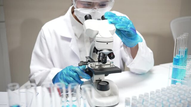 Focused scientist wearing mask, white coat and blue glove conducts research using microscope near laboratory glass test tube