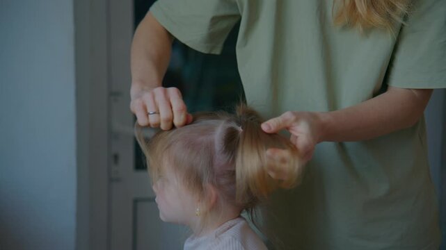 Caring adult gently styles a child's hair at home. The child stands still, trusting the adult's hands. Soft lighting creates a warm, intimate moment. Suitable for parenting, family care