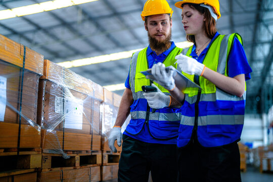 Male warehouse supervisor with a beard and his female coworker in protective workwear aim a glowing barcode reader at stacked cartons in a large fulfilment centre with rows of stacked.