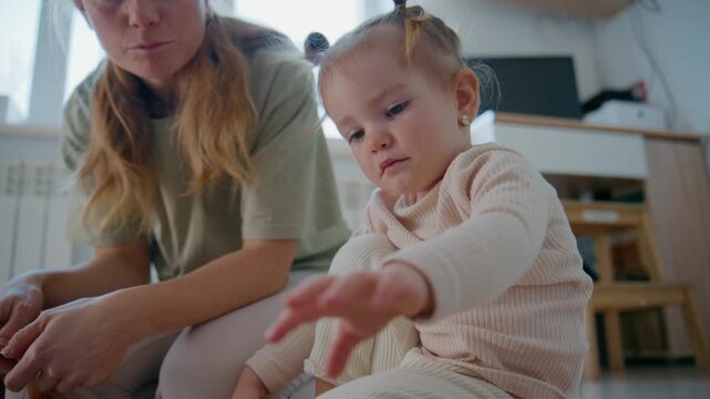 Young mother gently guides her toddler through playtime on the floor. The child focuses intently, reaching out with curiosity. The mother watches closely, offering calm support and encouragement