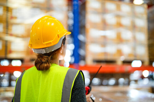 Asian female warehouse manager in a hard hat and yellow safety vest reads box labels stacked high on the racking beside tall warehouse racks loaded with packed cartons and goods with focus.
