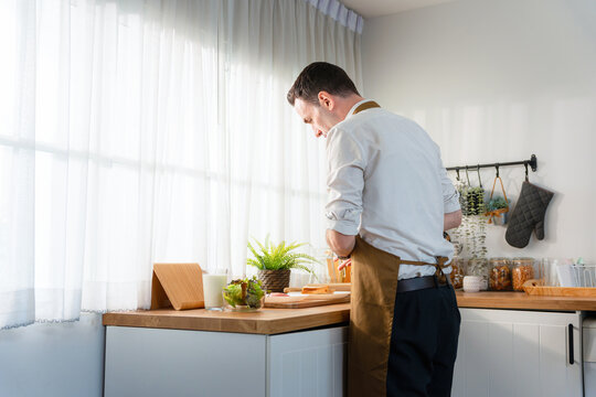 Man wearing apron preparing healthy breakfast in the kitchen, Caucasian man cooking in a bright modern home kitchen, Lifestyle portrait of a man making meal in the kitchen at home