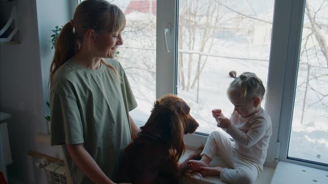 Mother and child sit by window with dog. Child feeds dog small treat. Mother smiles gently watching. Dog looks up calmly accepting food. Scene shows peaceful family moment indoors