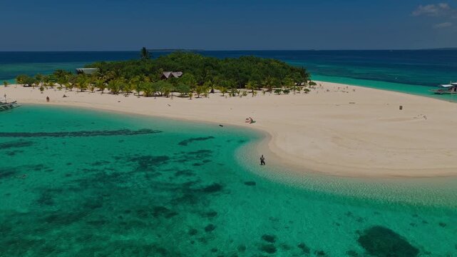 Aerial drone descent to land at Patawan Island tracking a man standing in crystal clear water, Balabac, Philippines. Inbound landing approach over shallow turquoise sea towards tropical beach.
