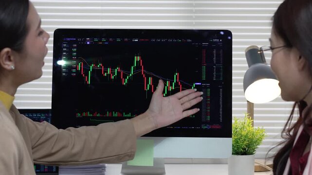 Focused woman trader and colleague analyzing stock investment chart on computer screen inside corporate office