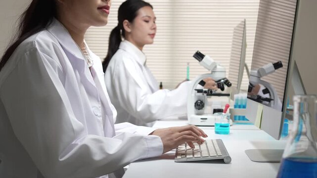Happy young Asian woman scientist conducting research in laboratory typing on computer keyboard near microscope