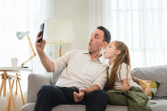 Cheerful father and young daughter taking a selfie with smartphone on sofa at home, Smiling dad and daughter bonding and looking at smartphone screen on couch in living room