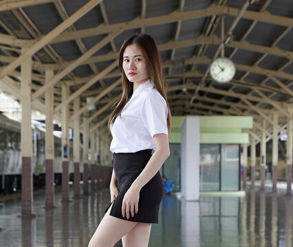 A young Asian woman in a white student uniform shirt and black skirt poses on a railway station platform