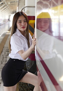 A young Asian woman in a white uniform shirt and black skirt steps up onto a train carriage at a station platform