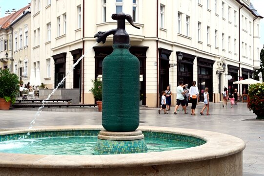 soda syphon bottle sculpture and fountain in public square. streetscape in Gyor, Hungary. layered green glass. shallow blue pool. popular landmark. outdoors, travel, tourism concept. people walking by