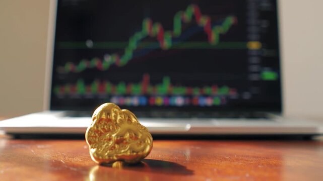 Gold nugget on wooden desk with stock market chart on laptop screen background