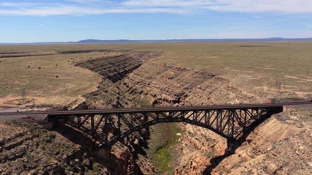 4K UHD drone flight showcasing a massive steel arch railroad bridge crossing a rugged canyon. The tracks stretch across the vast, flat Arizona desert landscape over a deep, dry river gorge.