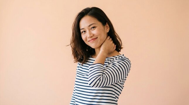 A young woman with dark hair wearing a striped shirt smiles while rubbing her neck in a studio setting.