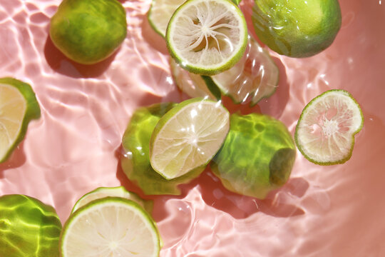 Top view of fresh whole green limes and slices floating in clear rippling water with a pink background.