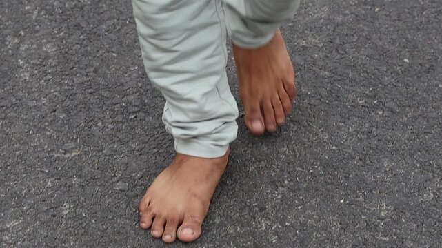 Close-up low angle view of an unrecognized person walking barefoot on a rough asphalt road towards the camera.
