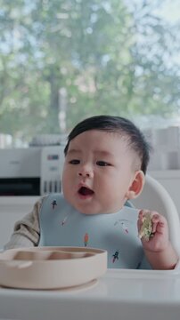 Adorable little baby toddler practicing baby-led weaning BLW at home with green bokeh background.