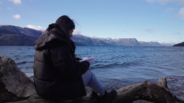 Woman reads beside Lake Traful in Villa Traful, Neuqu&eacute;n, Argentina, surrounded by crystal-clear water, driftwood, snow-capped Andes and tranquil Patagonian wilderness, static lakeside shot.