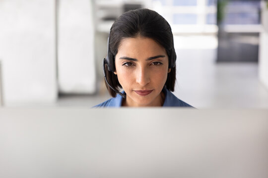 Closeup of female support agent in headset assisting client remotely, looks at computer monitor, listens attentively, providing professional customer or technical support services. Helpdesk workflow