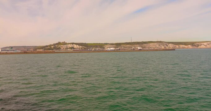Wide shot of the White Cliffs of Dover and harbor structures viewed across calm green sea under a hazy sky.