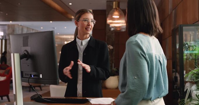 Pretty woman receptionist dressed in formal attire stands at reception front desk and serves female client, making check-in, explain hotel check-out rules, provide helpful information. Guests services
