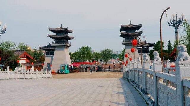 A wide static shot from a carved stone bridge overlooking the main entrance plaza of the garden, flanked by two towering traditional multi-tiered gray pillars.