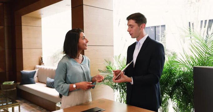 Male receptionist dressed in suit, stands at reception desk holding tablet, assisting to female guest, explaining amenities, friendly providing guidance about facilities. Hotel services, modern tech