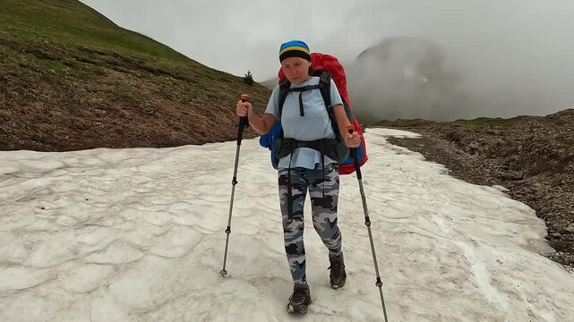 Female hiker walking in medium shot across mountain snow in Georgia, using trekking poles and backpack on alpine route for winter trekking, outdoor endurance, glacier travel, and adventure tourism
