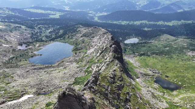 High-angle aerial drone view of a majestic mountain range with sharp rocky peaks, lush green pine forests, and pristine alpine lakes under a dramatic sky with scattered clouds