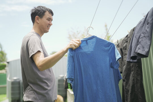 laundry, man, hanging, outdoor, drying, clothesline, chores: A middle-aged Asian man hangs a blue t-shirt to air dry in his backyard on a bright sunny day, promoting sustainable living.