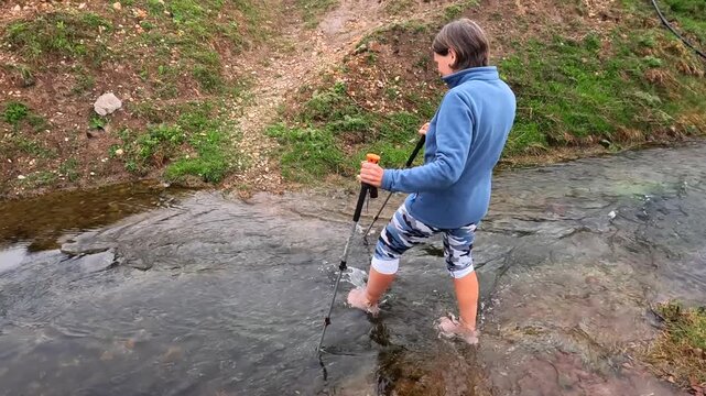 Woman crossing mountain stream side view in Georgia, walking barefoot through cold water with trekking poles on rural hiking route for travel, river safety, trekking tourism, and expedition footage