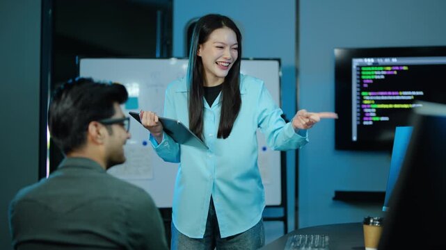 Smiling Asian female IT project manager holding a tablet and confidently explaining a new coding strategy to a developer during a late night brainstorming meeting in a modern software office.
