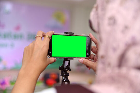 over-the-shoulder shot of a person holding a smartphone mounted on a tripod with a bright green chroma key screen, recording or streaming an event.