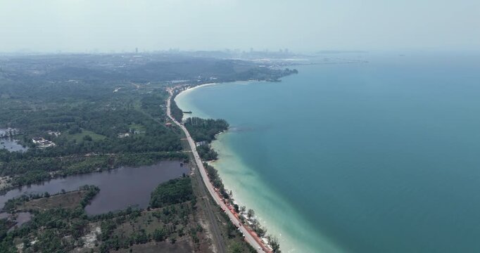 Aerial view: a coastal road stretches alongside a long, curved sandy beach, separating a lush green landscape with a dark pond from the sea with the Sihanoukville city skyline visible on the horizon.