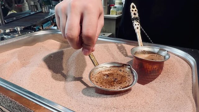 Close up of delicious oriental coffee boiling in copper pots placed in hot sand container. Professional barista hand holding ibrik handle making traditional turkish espresso beverage outdoors.