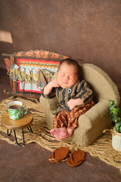 newborn baby boy sleeping in traditional javanese lurik and jarik clothes pose on mini sofa like an old man drinking tea, in vertical format.