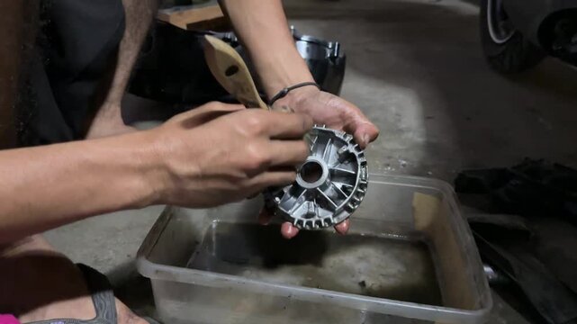 Close up of a mechanic cleaning a motorcycle CVT pulley component with a brush and solvent in a workshop.