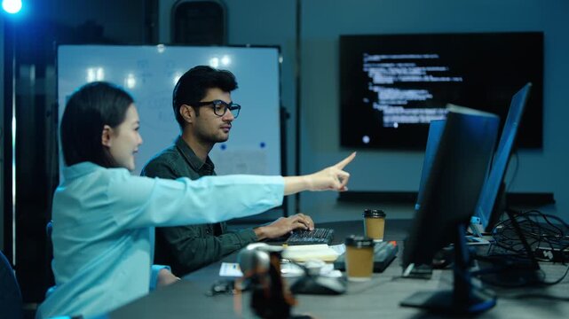 Diverse tech professionals collaborate on complex software development in a dark office. Asian female programmer points at a computer screen with code while a male developer types late at night.